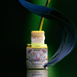 May include: A yellow and white candle sits on a decorative box with the text "MORFOSIA" and "SAFF & Co." The box features a floral design with a central illustration. A blue and green feather and a yellow stem are in the background.