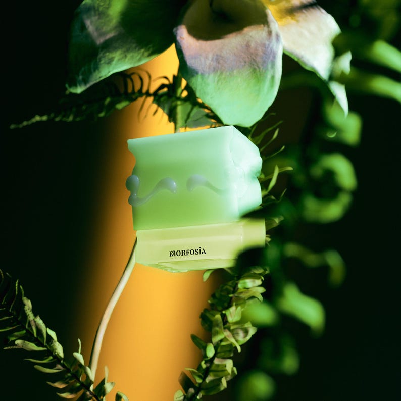 May include: A pale green soap bar with a unique, uneven shape and white decorative details. It rests on a light yellow rectangular object bearing the word "MORFOSIA". The image is set against a backdrop of green foliage and a yellow background.