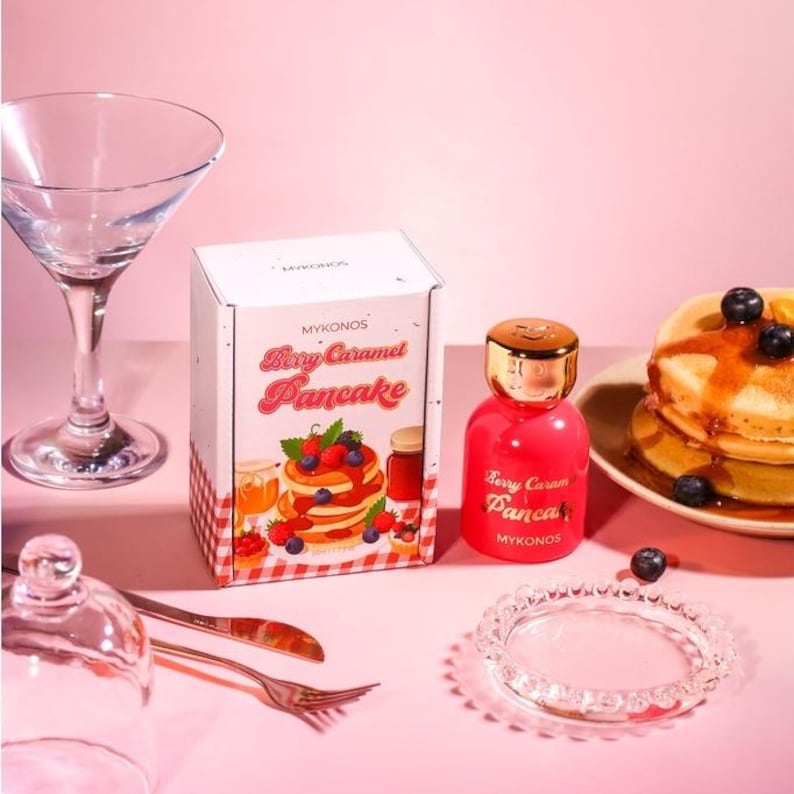 Puede incluir: Imagen de producto con tem&aacute;tica rosa que presenta un frasco de perfume y una caja con la etiqueta "Berry Caramel Pancake" con una ilustraci&oacute;n de panqueques. Una pila de panqueques con ar&aacute;ndanos y jarabe est&aacute; en un plato. Tambi&eacute;n hay una copa de martini y cubiertos.