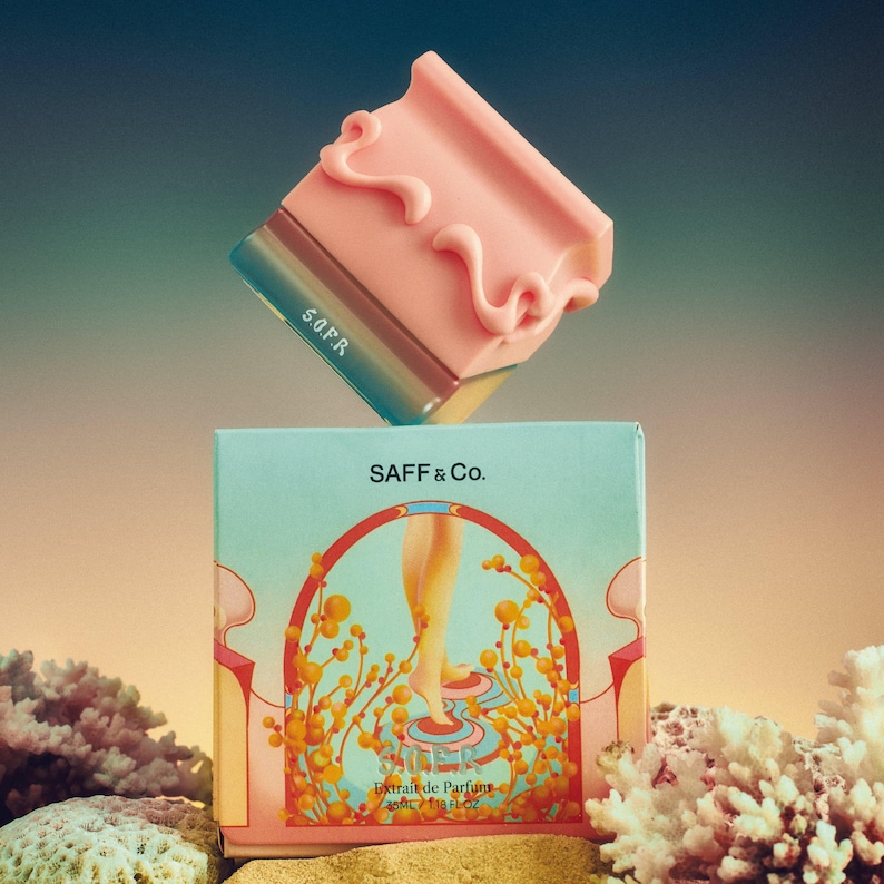 May include: A pink perfume bottle with a decorative top and the text "S.O.F.R." sits above a box with the text "SAFF & Co." and an illustration of a person's legs. The box also has the text "Extrait de Parfum 15ML / 0.71fl oz."