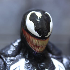 May include: Close-up of a black and white Venom action figure with a menacing grin. The figure has a detailed sculpt and paint job, with a white symbiote design on the face.