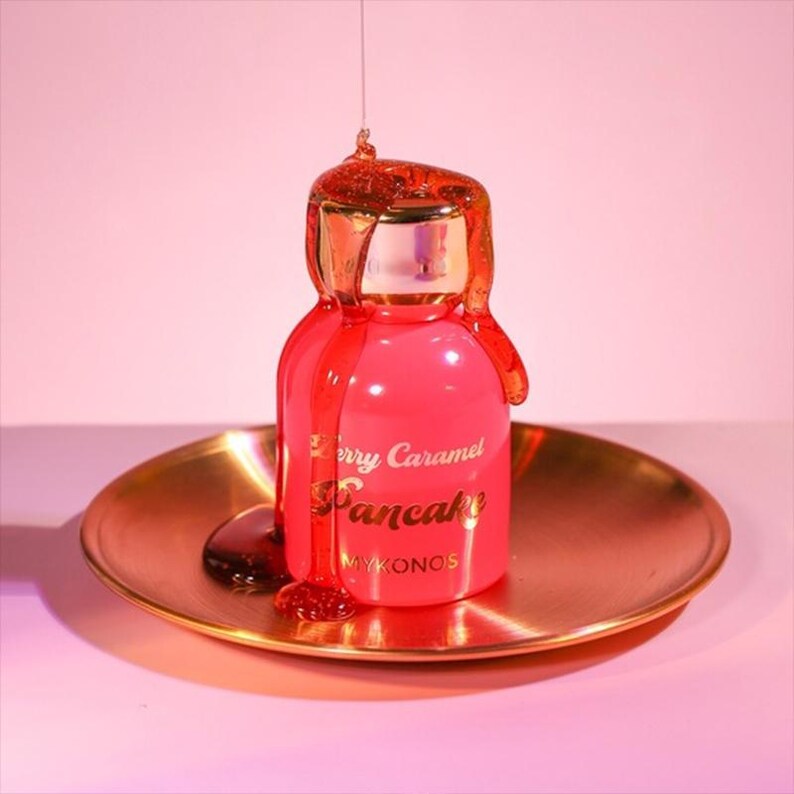 Puede incluir: Una botella rosa con las palabras "Berry Caramel Pancake" en letras doradas, con una tapa dorada y un plato dorado. Un l&iacute;quido viscoso de color &aacute;mbar gotea de la botella, creando un efecto visualmente atractivo. La palabra "Mykonos" tambi&eacute;n es visible.