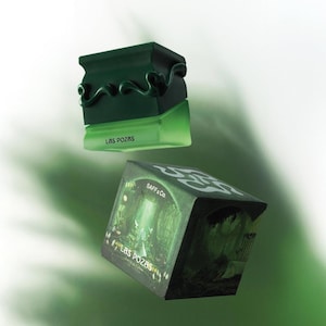 May include: A dark green perfume bottle with a decorative top and a matching box. The bottle has the text "LAS POZAS" on the front. The box features a forest scene and the text "SAFF & Co."