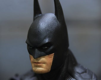May include: Close-up of a black Batman mask with a detailed texture. The mask covers the face of a figure, revealing only the eyes and a portion of the chin.