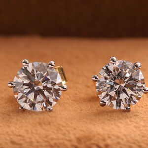 May include: A pair of round diamond stud earrings set in a silver metal setting. The earrings are on a brown surface.