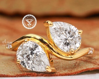 May include: A gold ring with two pear-shaped diamonds set in a twisted band. The diamonds are set in a prong setting and are surrounded by smaller diamonds. The ring is on a brown background.