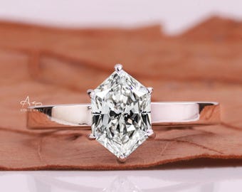 May include: A silver ring with a large, clear, hexagonal-cut diamond as the centerpiece. The diamond is set on a simple, polished silver band. The ring is displayed on a brown surface, reflecting light.