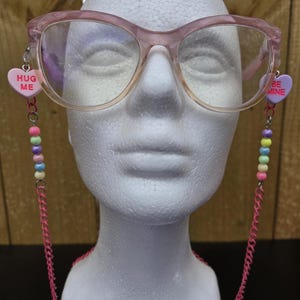 May include: Pink cat-eye glasses with a pink chain and colorful beads. The chain has a pink heart charm on each end that says "Hug Me" and "Be Mine".
