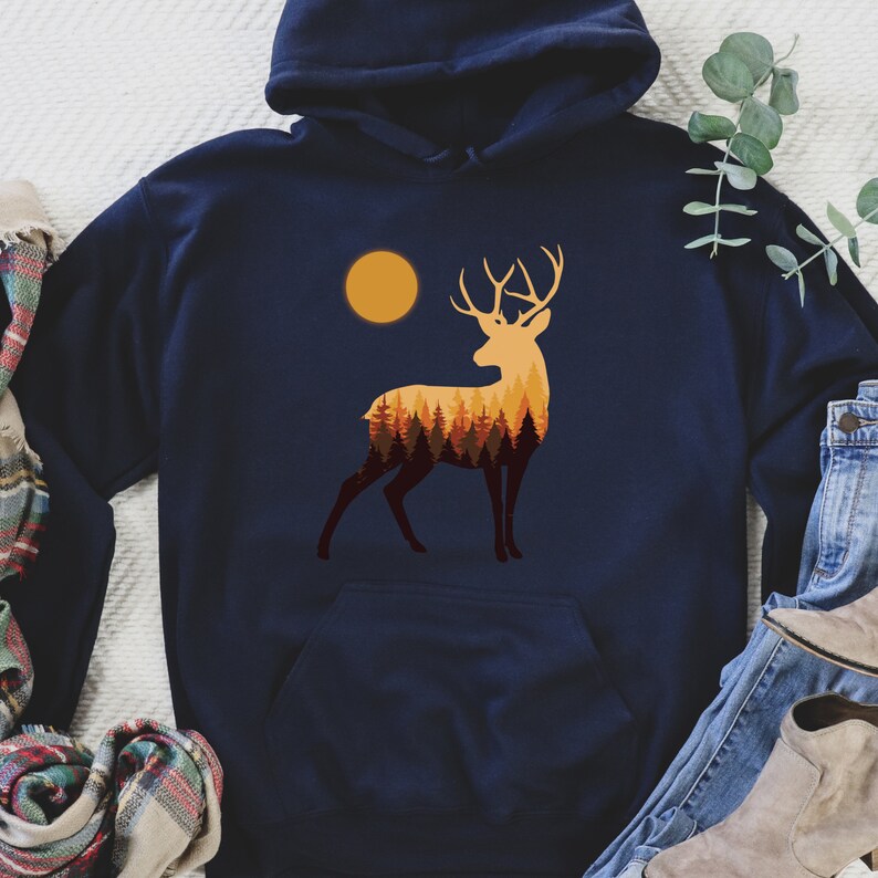 May include: A navy blue hoodie with a graphic of a deer silhouette with a forest inside. The deer is standing in front of a yellow sun.
