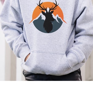 Puede incluir: Sudadera con capucha gris claro con bolsillo de canguro. Presenta un gr&aacute;fico circular de una silueta de ciervo con cuernos, sobre un fondo de monta&ntilde;as naranjas y azules. El texto "Outdoorsygoods online" est&aacute; en la parte inferior.