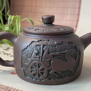 May include: A dark brown clay teapot featuring a relief carving of a horse-drawn carriage on its side. The lid is decorated with a geometric Greek key pattern around the edge and has a small knob handle. The teapot has a curved handle and spout.