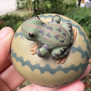 May include: A ceramic frog figurine in shades of green, brown, and black, resting on a round, light-colored base with a wavy, dark green pattern. The frog has heart-shaped spots and orange feet.