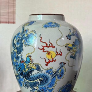 May include: A white ceramic vase with a wide body and a narrow neck. The vase is decorated with blue dragons, clouds, and a yellow sun with red flames. The rim is brown.