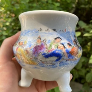 May include: A white ceramic incense burner with three short legs. The exterior features a colorful illustration of figures in a landscape with text in Chinese characters. The rim has a flared shape.