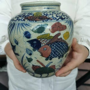 May include: A ceramic vase with a cream-colored base and blue, red, yellow, and green painted details. The vase features a design of fish and clouds. The vase is held by a person.