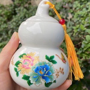 May include: White ceramic gourd-shaped container with a lid, adorned with floral and butterfly designs in pink, blue, and gold. It has a yellow tassel and a red bead attached to the top. The container is held in a hand.