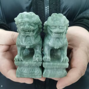 May include: Two jade Foo Dog statues. The statues are a muted green color, carved with detailed features, including a lion-like face, and seated on a decorative base. The Foo Dogs are held in a person's hands.