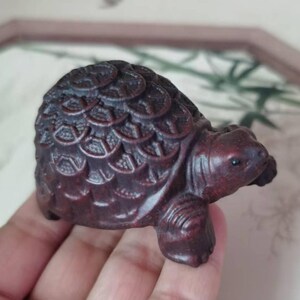 May include: A small, intricately carved wooden turtle figurine. The dark reddish-brown turtle has detailed shell patterns and a smooth, rounded head. The figurine is held in a hand, showcasing its size and craftsmanship.