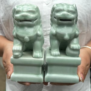 May include: Two celadon-colored ceramic guardian lion statues. Each lion is seated on a rectangular base, with detailed features including a mane, paws, and a smiling expression. The statues are held in front of a white background.
