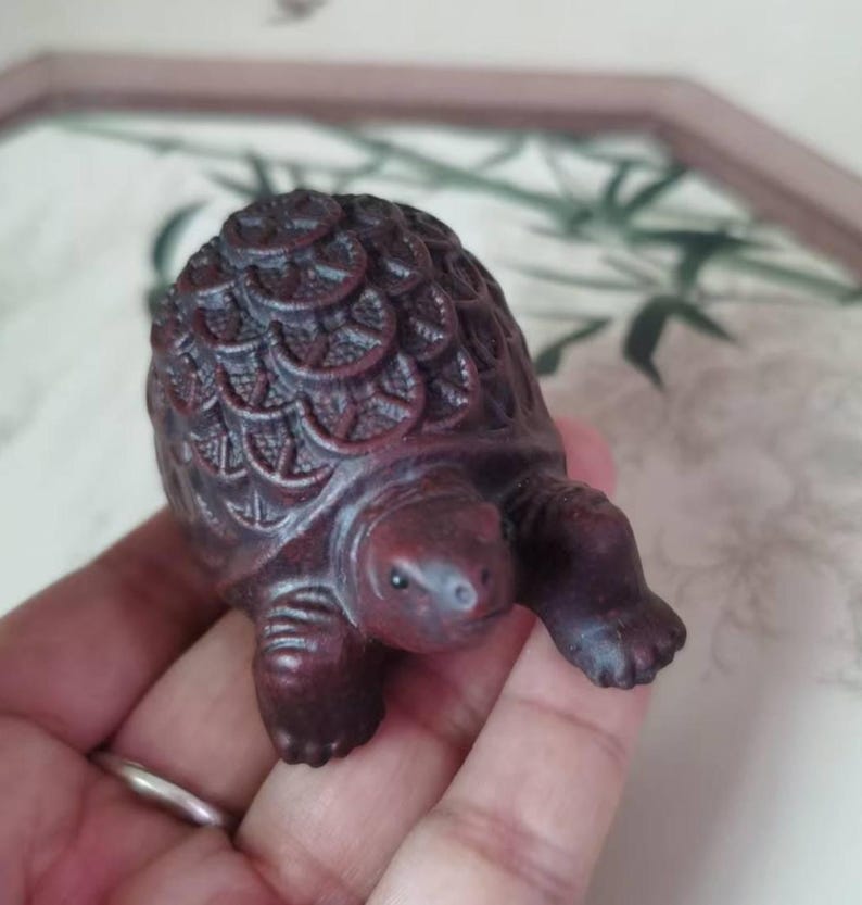 May include: A small, intricately carved wooden turtle figurine. The dark reddish-brown turtle has detailed shell patterns and is posed with its legs extended. The figurine is held in a hand.