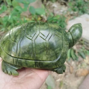 May include: A carved green stone turtle figurine. The turtle has a detailed shell pattern and is held in a hand. The figurine is a decorative item, likely for display or collection. The stone has a mottled appearance.