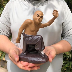 May include: A ceramic figurine of a bald, muscular figure in a martial arts pose. The figure is brown-skinned with a stern expression, wearing dark brown pants and a matching base. The figurine is held in a person's hands.