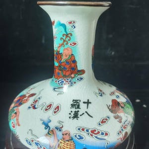 May include: A white ceramic vase with a bulbous body and a flared neck. The vase is decorated with colorful illustrations of figures and clouds, with Chinese characters. The rim is brown.
