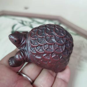 May include: A small, reddish-brown turtle figurine. The turtle has a detailed shell pattern and small, dark eyes. The figurine is held in a hand, showcasing its size and craftsmanship. This decorative item could be a collectible or a unique home accent.