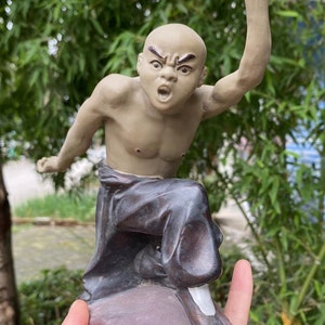 May include: A tan-colored figurine of a bald martial artist in a fighting stance. The figure is wearing dark pants and white shoes, with one arm raised and mouth open in a yell. The figurine is on a purple base.