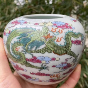 May include: A white ceramic vase with a detailed dragon design. The dragon is green, yellow, and blue, set against a backdrop of colorful clouds. The vase has a round shape and a wide opening, suitable for decorative use.