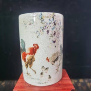 May include: A cylindrical ceramic vase with a crackle glaze finish. The vase features a hand-painted design of a rooster, hen, and chicks, with floral accents in shades of red, brown, blue, and purple. The vase is displayed on a red wooden surface.