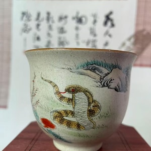 May include: A ceramic teacup with a hand-painted design of a snake with a red tongue and eyes. The cup has a speckled, off-white background with a brown rim and is set on a wooden surface. The design includes green grass and a red flower.