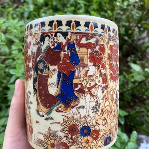 May include: Cream-colored ceramic cylinder featuring a Japanese-style illustration. The design includes figures in traditional attire, floral patterns, and architectural elements in red, blue, and brown hues. The cylinder has a slightly aged appearance.
