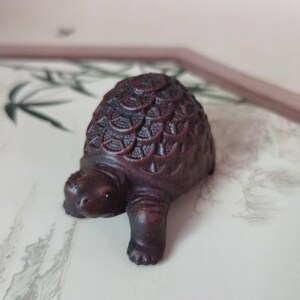 May include: A small, intricately carved wooden turtle figurine in shades of dark brown. The turtle's shell features a detailed, textured pattern. The figurine is positioned on a patterned surface, suggesting a decorative or collectible item.