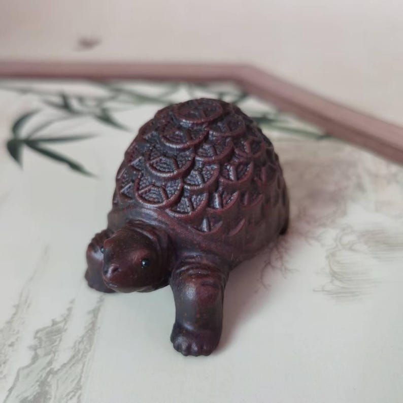 May include: A small, intricately carved wooden turtle figurine in shades of dark brown. The turtle's shell features a detailed, textured pattern. The figurine is positioned on a patterned surface, suggesting a decorative or collectible item.