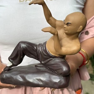 May include: A ceramic figurine of a bald person in a martial arts stance, featuring a tan-colored upper body and dark brown trousers. The figure is posed in a low crouch on a dark brown, rock-like base, with arms raised and hands positioned forward.