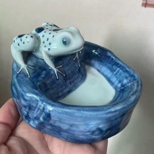 May include: A ceramic frog figurine with a blue and white spotted pattern, perched on a blue dish. The dish has a textured, wave-like design. The frog has a light blue hue with dark blue spots.