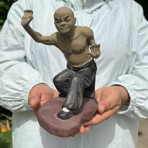 May include: A ceramic figurine of a bald martial artist in a fighting stance. The figure is wearing dark pants and black shoes, with a muscular upper body. It is mounted on a reddish-brown base.