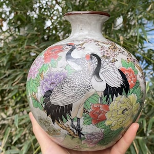 May include: A white ceramic vase with a round body and a short neck. The vase is decorated with a floral pattern and two cranes in black, white, and gray.