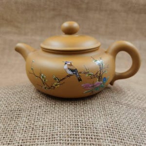 May include: A tan ceramic teapot with a rounded body, a curved handle, and a spout. The lid has a small knob. A painted bird and floral design decorates the teapot. The teapot is sitting on a burlap surface.
