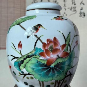 May include: A white ceramic jar with a lid, featuring a hand-painted design of a large pink lotus flower, green lily pads, and two small birds perched on stems. The jar has a rounded body and a smooth, glossy finish with detailed botanical art.