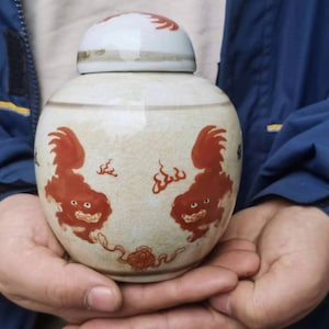 May include: A cream-colored ceramic jar with a matching lid. The jar is decorated with two red mythical creatures and other red accents. The jar is held in a person's hands.