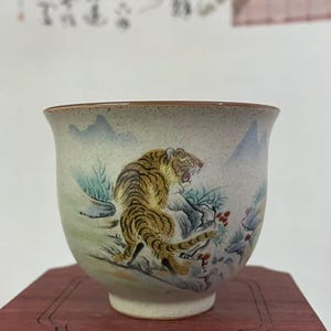 May include: A ceramic teacup with a detailed painting of a tiger in shades of yellow, brown, and black. The cup has a light green and beige background with mountain and plant details. The cup sits on a dark wooden stand.