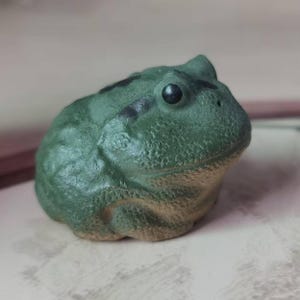 May include: A ceramic frog figurine in shades of green. The frog has a textured surface, black markings, and a dark eye. The figurine is a decorative item, possibly for a shelf or desk.