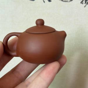 May include: A small, brown ceramic teapot with a rounded body, a curved handle, and a small spout. The lid has a small knob on top. The teapot is held in a hand, suggesting its size. The overall design is simple and elegant.