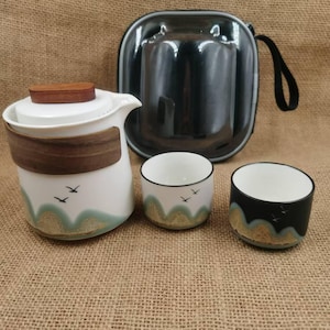 May include: A white and wood-toned tea set with a teapot, two cups, and a black carrying case. The teapot and cups feature a landscape design with flying birds. The cups are white and black.