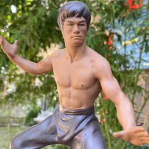 May include: A detailed figurine of a martial artist in a dynamic pose. The figure is shirtless, with defined musculature, and wearing dark gray pants. The statue has a focused expression, with arms outstretched, suggesting movement. The background is blurred with green foliage.