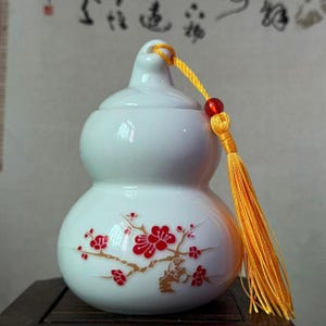 May include: A white ceramic gourd-shaped container with a lid, adorned with red floral branch designs. It features a yellow tassel and a red bead. The container is likely used for decorative or storage purposes.