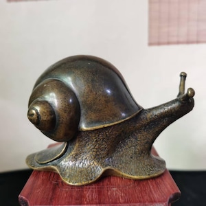 May include: A bronze snail sculpture with a detailed shell and textured body. The snail is posed on a small, reddish-brown wooden base, showcasing its antennae and overall form. The sculpture has a vintage aesthetic.