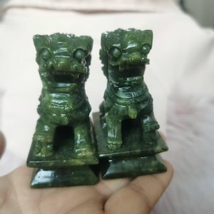 May include: Two jade green Foo Dog statues. The statues are carved with detailed features, including expressive eyes and ornate collars. Each dog sits atop a rectangular base. The statues are held in a hand.